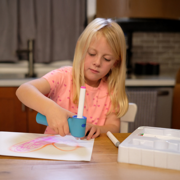 A young girl with blond hair is seen at a table in the kitchen using the airbrush sprayer on a piece of paper with a heart on it. The plastic mold for the sprayer from the kit is seen to the right on the table. One of the included dual-ended markers is seen in between the piece of paper and the plastic mold, but closer to the plastic mold.